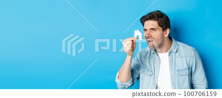 Real estate concept. Man looking at tiny house maket with confused face, standing puzzled against blue background 100706159
