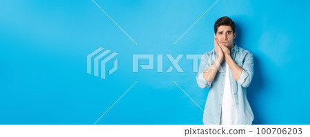 Portrait of man with toothache grimacing from pain and touching cheek, standing against blue background 100706203