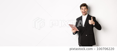 Portrait of handsome businessman in suit, showing thumb-up and making a compliment, praising something on digital tablet, standing over white background Portrait of handsome businessman in suit, showing thumb-up and making a compliment, praising something on digital tablet, standing over white background 100706220