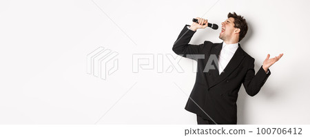Portrait of handsome man singing a song with passion, standing in black suit, holding microphone and performning, posing over white background 100706412