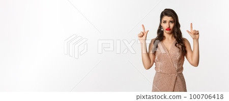New Year, christmas and celebration concept. Gloomy and sad glamour girl in party dress pointing fingers up, sulking and crying about something, standing over white background 100706418