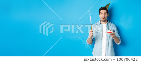 Happy young man celebrating birthday in party hat, holding b-day cake and smiling, standing over blue background 100706428