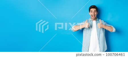 Smiling adult man showing thumbs up with excited face, like something awesome, approving product, standing against blue background 100706461