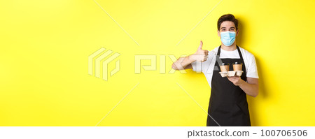 Concept of covid-19, cafe and social distancing. Young male barista in medical mask and black apron holding takeaway coffee cups, showing thumb up, yellow background Concept of covid-19, cafe and social distancing. Young male barista in medical mask and black apron holding takeaway coffee cups, showing thumb up, yellow background 100706506