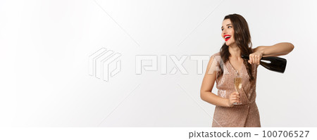 Winter holidays celebration concept. Excited woman pouring glass of champagne and talking to someone, looking left, having fun on party, standing in elegant dress Winter holidays celebration concept. Excited woman pouring glass of champagne and talking to someone, looking left, having fun on party, standing in elegant dress 100706527