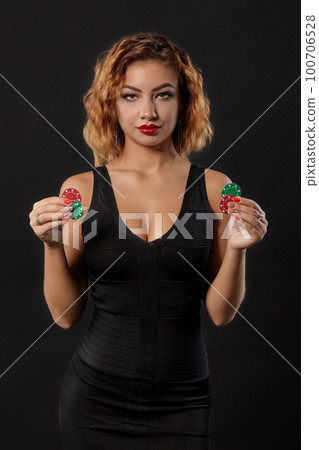 Ginger girl wearing dark dress is posing holding red and green chips in her hands standing against black studio background. Casino, poker. Close-up. Ginger girl wearing dark dress is posing holding red and green chips in her hands standing against black studio background. Casino, poker. Close-up. 100706528