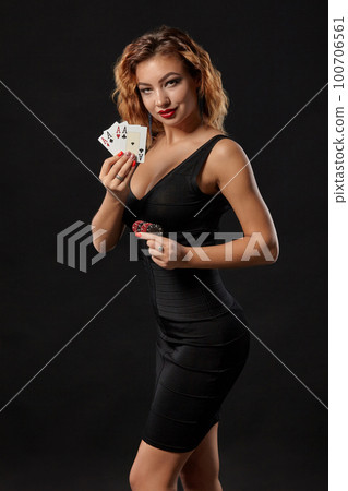 Ginger girl in a dark dress posing holding playing cards and chips in her hands standing against a black studio background. Casino, poker. Close-up. 100706561