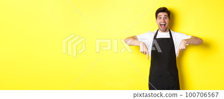 Waiter, coffee shop barista in black apron pointing fingers down, looking amazed, standing over yellow background 100706567