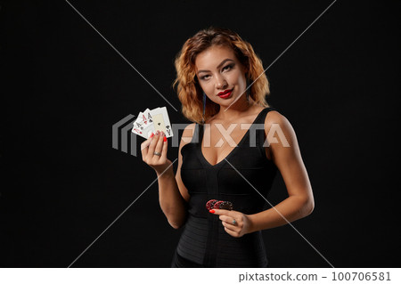 Ginger girl in a dark dress posing holding playing cards and chips in her hands standing against a black studio background. Casino, poker. Close-up. 100706581