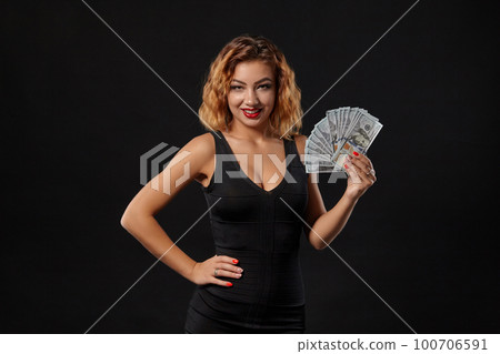 Ginger girl in dark dress posing holding fan of hundred-dollar bills in her hands standing against a black studio background. Casino, poker. Close-up. Ginger girl in dark dress posing holding fan of hundred-dollar bills in her hands standing against a black studio background. Casino, poker. Close-up. 100706591