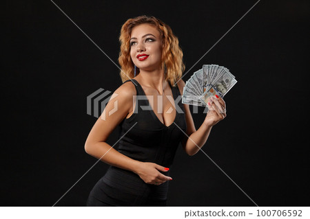 Ginger girl in dark dress posing holding fan of hundred-dollar bills in her hands standing against a black studio background. Casino, poker. Close-up. Ginger girl in dark dress posing holding fan of hundred-dollar bills in her hands standing against a black studio background. Casino, poker. Close-up. 100706592