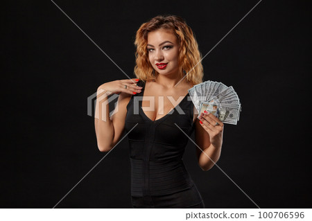 Ginger girl in dark dress posing holding fan of hundred-dollar bills in her hands standing against a black studio background. Casino, poker. Close-up. 100706596