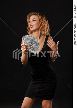 Ginger girl in dark dress posing holding fan of hundred-dollar bills in her hands standing against a black studio background. Casino, poker. Close-up. Ginger girl in dark dress posing holding fan of hundred-dollar bills in her hands standing against a black studio background. Casino, poker. Close-up. 100706606