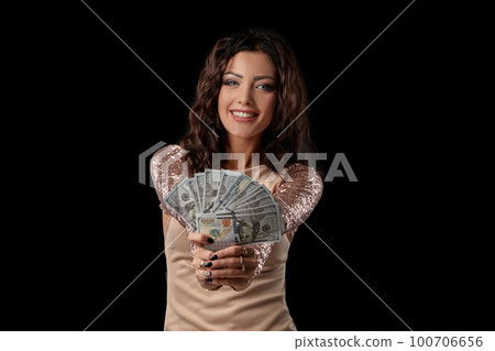 Brunette girl in shiny dress posing holding fan of hundred dollar bills in her hands standing on black studio background. Casino, poker. Close-up. 100706656