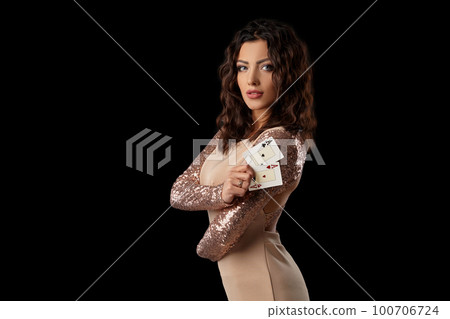 Brunette girl wearing shiny dress posing holding two playing cards in her hand standing against black studio background. Casino, poker. Close-up. 100706724