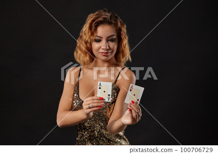Ginger girl wearing shiny dress is posing holding two playing cards in her hands standing against a black studio background. Casino, poker. Close-up. 100706729