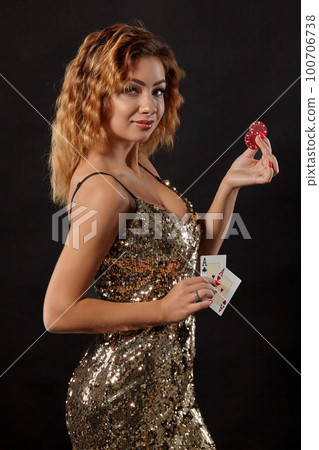 Ginger girl in shiny dress posing holding two playing cards and chips in her hands standing against black studio background. Casino, poker. Close-up. Ginger girl in shiny dress posing holding two playing cards and chips in her hands standing against black studio background. Casino, poker. Close-up. 100706738