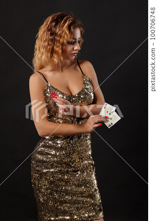 Ginger girl in shiny dress posing holding two playing cards and chips in her hands standing against black studio background. Casino, poker. Close-up. 100706748