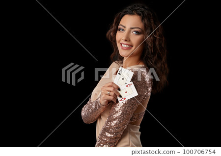 Brunette girl wearing shiny dress posing holding two playing cards in her hand standing against black studio background. Casino, poker. Close-up. Brunette girl wearing shiny dress posing holding two playing cards in her hand standing against black studio background. Casino, poker. Close-up. 100706754