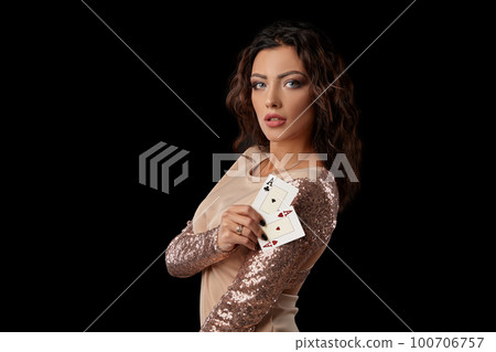Brunette girl wearing shiny dress posing holding two playing cards in her hand standing against black studio background. Casino, poker. Close-up. Brunette girl wearing shiny dress posing holding two playing cards in her hand standing against black studio background. Casino, poker. Close-up. 100706757