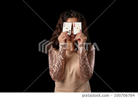 Brunette girl wearing shiny dress posing holding two playing cards in her hands standing against black studio background. Casino, poker. Close-up. Brunette girl wearing shiny dress posing holding two playing cards in her hands standing against black studio background. Casino, poker. Close-up. 100706764