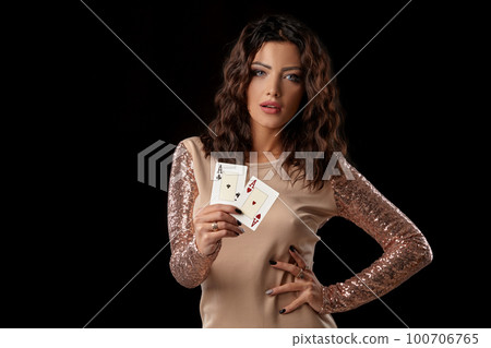 Brunette girl wearing shiny dress posing holding two playing cards in her hand standing against black studio background. Casino, poker. Close-up. Brunette girl wearing shiny dress posing holding two playing cards in her hand standing against black studio background. Casino, poker. Close-up. 100706765