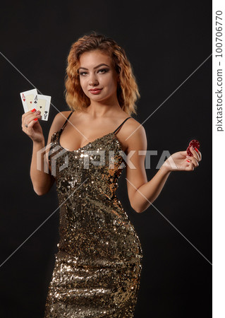 Ginger girl in shiny dress posing holding two playing cards and chips in her hands standing against black studio background. Casino, poker. Close-up. Ginger girl in shiny dress posing holding two playing cards and chips in her hands standing against black studio background. Casino, poker. Close-up. 100706770