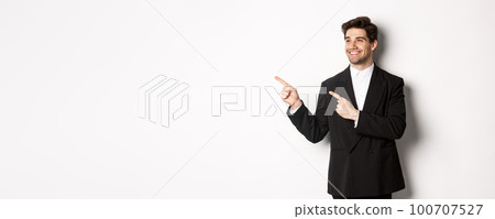 Portrait of successful handsome man in suit, pointing and looking left with pleased smile, showing promo banner, standing over white background 100707527