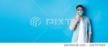 Concept of covid-19, social distancing and quarantine. Young man in medical mask searching for something, looking through magnifying glass, standing against blue background 100707684
