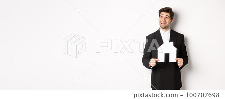 Image of handsome investor in black suit, holding house maket and looking at upper left corner thoughtful, standing against white background 100707698