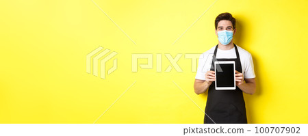 Concept of covid-19, small business and pandemic. Smiling barista in medical mask showing tablet screen, standing over yellow background Concept of covid-19, small business and pandemic. Smiling barista in medical mask showing tablet screen, standing over yellow background 100707902