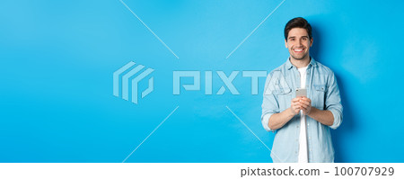 Handsome bearded man in casual outfit smiling at camera, checking smartphone, standing against blue background Handsome bearded man in casual outfit smiling at camera, checking smartphone, standing against blue background 100707929