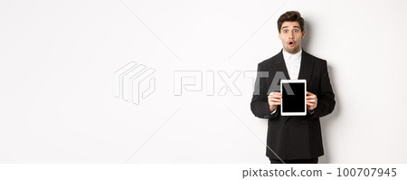 Image of surprised handsome man in black suit, showing digital tablet screen and looking amazed, standing against white background Image of surprised handsome man in black suit, showing digital tablet screen and looking amazed, standing against white background 100707945