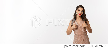 Concept of celebration, holidays and party. Beautiful female model with red lips, wearing glamour dress and showing thumbs up in approval, like and agree, praise good choice 100707970