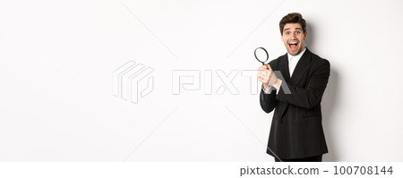 Handsome businessman in black suit, holding magnifying glass and smiling, found something, standing against white background 100708144