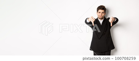 Portrait of skeptical and disappointed man in black suit, frowning upset, showing thumbs-down, dislike something bad, standing over white background 100708259
