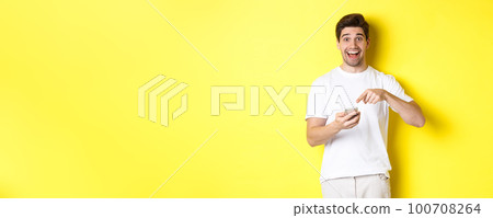 Happy man pointing at smartphone to show promo, check out internet offer, standing over yellow background 100708264