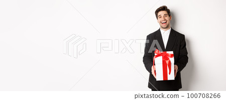 Concept of christmas holidays, celebration and lifestyle. Joyful handsome man in black suit, holding xmas gift and smiling, standing against white background 100708266