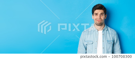 Close-up of skeptical and awkward guy smirking, feeling uncomfortable, standing over blue background 100708300