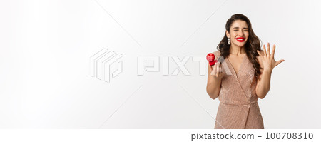 Excited young woman in glamour dress being engaged, showing finger with ring and looking happy, receive marriage proposal, standing over white background Excited young woman in glamour dress being engaged, showing finger with ring and looking happy, receive marriage proposal, standing over white background 100708310