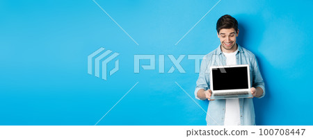 Excited handsome man looking at screen, showing promo offer on computer screen, smiling amazed, standing over blue background 100708447