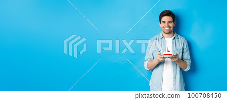 Smiling man celebrating birthday, holding b-day cake with candle, standing over blue background 100708450