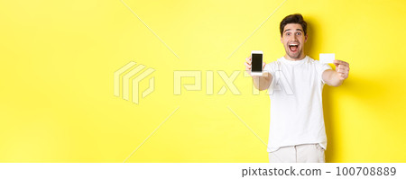 Handsome caucasian man showing smartphone screen and credit card, concept of mobile banking and online shopping, yellow background 100708889