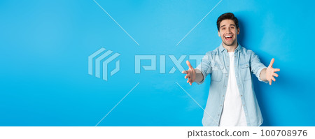 Friendly man stretching hands forward and welcome you, greeting guests or reaching for hugs, standing against blue background 100708976