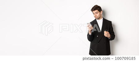 Portrait of thoughtful handsome businessman, drinking coffee and browsing in internet, looking at smartphone screen, standing against white background Portrait of thoughtful handsome businessman, drinking coffee and browsing in internet, looking at smartphone screen, standing against white background 100709185