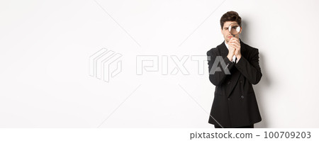 Image of serious businessman in black trendy suit, looking through magnifying glass, looking for employees, standing against white background 100709203