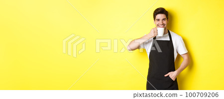 Cafe barista drinking cup of coffee and smiling, wearing black apron, standing over yellow background 100709206