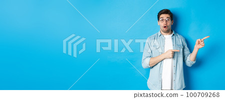 Surprised man in glasses pointing right at copy space, showing promo offer on blue background, standing over blue background 100709268