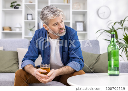 Close up of adult mature man lonely depressed drinking strong alcohol sitting on sofa in living room on day. 100709519