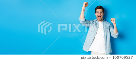Excited handsome man triumphing, raising hands up and shouting for joy, celebrating victory, standing against blue background Excited handsome man triumphing, raising hands up and shouting for joy, celebrating victory, standing against blue background 100709527